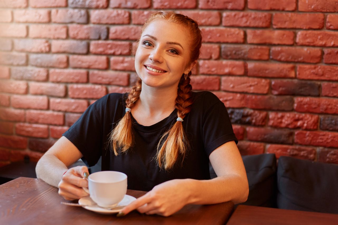 Femme d'une trentaine d'années aux cheveux bouclés roux, écharpe colorée, sourire chaleureux, photographiée dans un café lumineux
