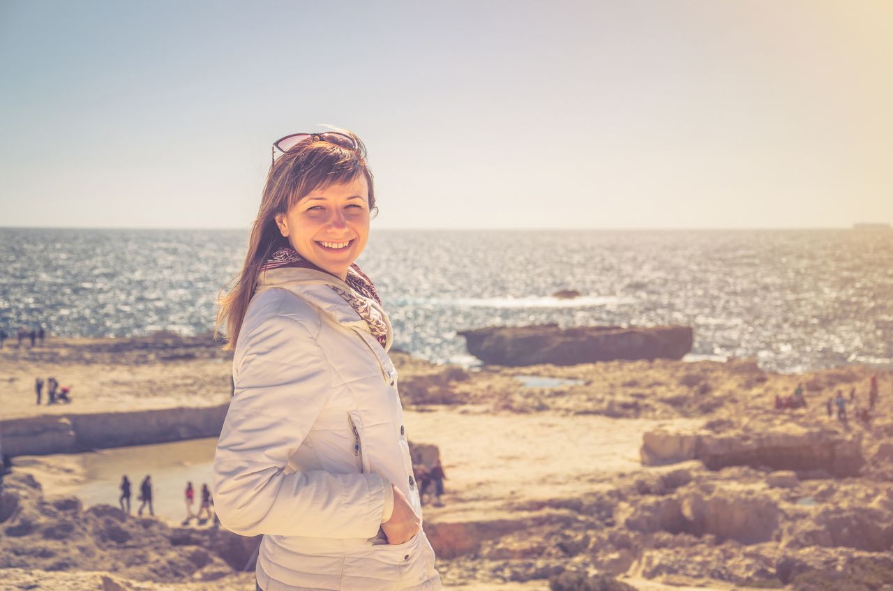 Jeune femme d'une trentaine d'années aux cheveux châtains attachés, coupe-vent outdoor, sourire franc, photographiée sur un sentier côtier face à la mer